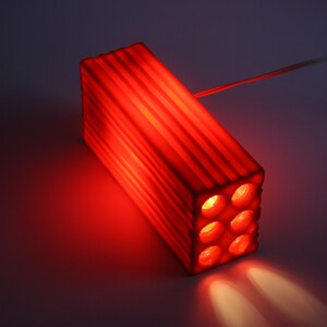 Pu&ograve; includere: Un apparecchio di illuminazione rettangolare rosso con sei aperture circolari. L'apparecchio ha una struttura a coste sui lati ed emette un bagliore rosso brillante. La luce brilla verso il basso, creando un effetto riflettore.