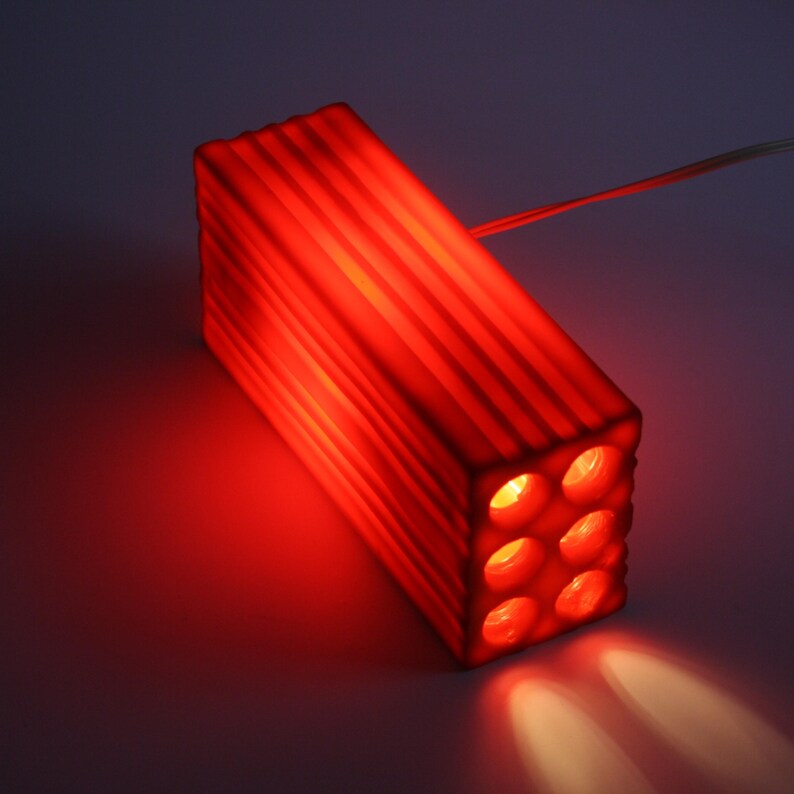 Pu&ograve; includere: Un apparecchio di illuminazione rettangolare rosso con sei aperture circolari. L'apparecchio ha una struttura a coste sui lati ed emette un bagliore rosso brillante. La luce brilla verso il basso, creando un effetto riflettore.