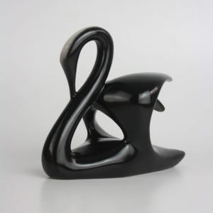 Puede incluir: Una escultura de cerámica negra lisa y brillante de un cisne con cuello curvado y cuerpo estilizado. La figura presenta una forma fluida y continua con un cuello arqueado y una espalda redondeada, mostrando un acabado oscuro y sólido.