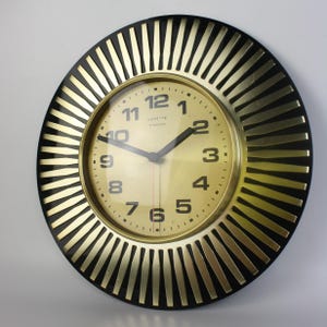 May include: A round wall clock featuring a metallic gold face with black Arabic numerals and black hands. The clock is surrounded by a sunburst-style frame with alternating black and gold rays. The text Vedette Transistor is printed on the face.
