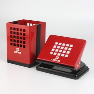 Puede incluir: Juego de escritorio rojo que incluye un portalápices y una perforadora. El portalápices tiene forma cuadrada con un diseño de rejilla. La perforadora es cuadrada con un patrón de rejilla y la palabra "CHECKER" impresa.