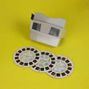 Może przedstawiać: Biały, zabytkowy wizjer View-Master z trzema okrągłymi rolkami obrazów. Wizjer ma dwa okulary i dźwignię. Rolki obrazów są białe z małymi kwadratowymi obrazami i tekstem. Tło jest żółte.