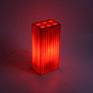 Pu&ograve; includere: Una lampada rossa rettangolare con esterno a coste e aperture circolari sulla parte superiore. La lampada &egrave; illuminata, emettendo un bagliore rosso caldo. Un sottile cavo bianco si estende dalla base.