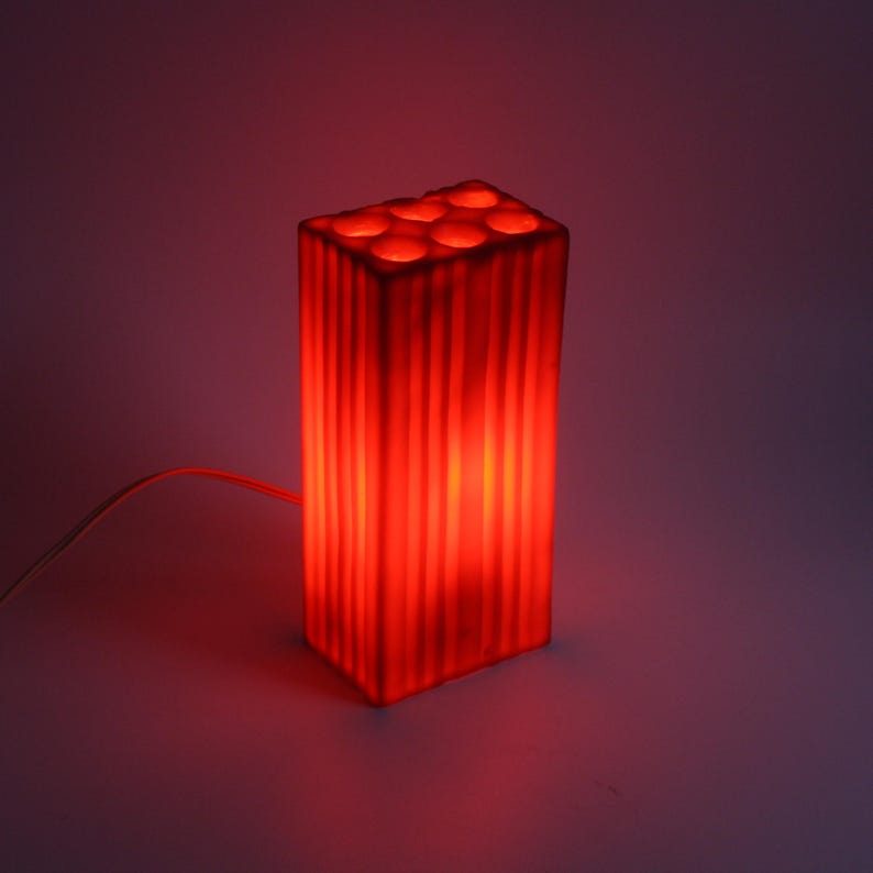 Pu&ograve; includere: Una lampada rossa rettangolare con esterno a coste e aperture circolari sulla parte superiore. La lampada &egrave; illuminata, emettendo un bagliore rosso caldo. Un sottile cavo bianco si estende dalla base.