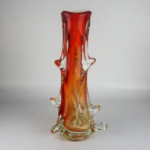 May include: A tall, decorative glass vase with a vibrant gradient of colors. The vase transitions from a clear base to shades of amber and orange, culminating in a rich red at the top. It features unique, organically shaped protrusions along its sides.