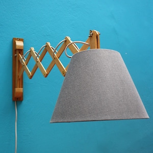 Peut inclure: Une lampe murale dotée d'un bras articulé en accordéon en bois clair qui s'étend vers l'extérieur. La lampe est surmontée d'un abat-jour conique en tissu gris texturé et comprend un cordon d'alimentation blanc suspendu à la base.