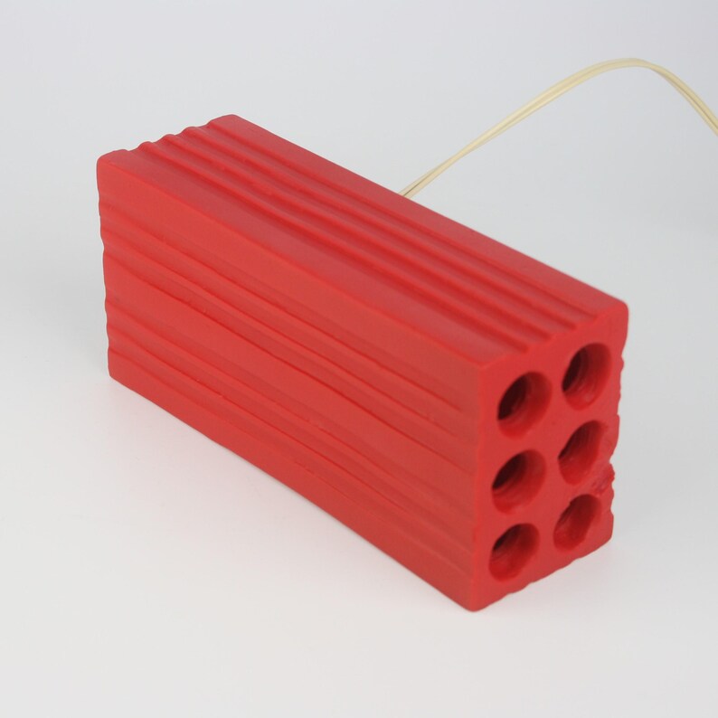 Pu&ograve; includere: Un oggetto rettangolare rosso brillante con sei fori circolari e una superficie scanalata. Un cavo di colore chiaro si estende dall'oggetto. Probabilmente si tratta di un oggetto decorativo o funzionale, come un supporto o una base.