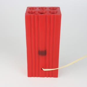 Pu&ograve; includere: Un blocco rettangolare rosso brillante con sei fori circolari sulla parte superiore e scanalature verticali sui lati. Un sottile cordino bianco sporco esce da un foro vicino alla base. Il blocco sembra essere fatto di un materiale gommoso.