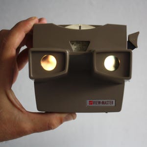 Könnte beinhalten: Ein Vintage View-Master-Betrachter in einem beigen Farbton, gehalten von einer Hand. Der Betrachter hat zwei Okulare mit beleuchteten Kreisen. Das View-Master-Logo ist auf der Vorderseite sichtbar. Ein klassisches Spielzeug.