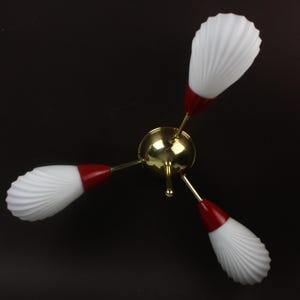 May include: A three-arm ceiling light fixture featuring a central polished gold-tone metal sphere. Each arm extends to a red conical base supporting a white, ribbed, shell-shaped glass shade. The design radiates outward from the center point.