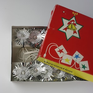 Puede incluir: Un juego de luces navideñas vintage con luces plateadas de oropel en forma de estrella en una caja de cartón. La caja roja presenta una estrella con campanas y la palabra "Decoration". Las luces están en un cable blanco.