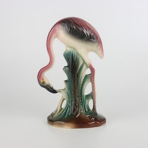 Puede incluir: Figura de flamenco de cerámica con cuello largo y curvado y cabeza mirando hacia abajo. El flamenco tiene colores rosa, negro y blanco, y se apoya sobre una base con detalles verdes y marrones. Es un objeto decorativo.