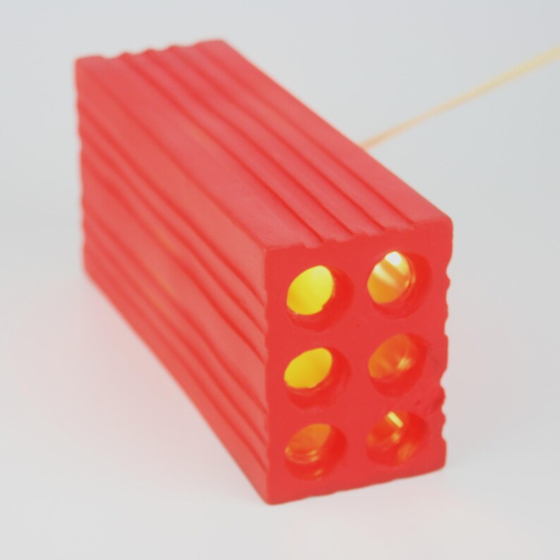 Pu&ograve; includere: Un blocco rettangolare rosso brillante con sei fori circolari. Il blocco presenta una texture a coste sulla parte superiore e sui lati. I fori rivelano un bagliore giallo caldo, suggerendo una fonte di luce interna. Probabilmente &egrave; una lampada decorativa.