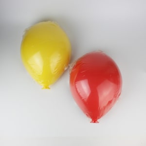 May include: Two balloon-shaped objects, one yellow and one red, are wrapped in clear plastic. The balloons have a smooth, glossy surface and are positioned against a white background. The yellow balloon is on the left, and the red balloon is on the right.