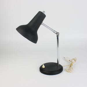 May include: A black desk lamp with a textured shade and a chrome-colored adjustable arm. The lamp has a round base with a small on/off switch and a white power cord. The lamp is designed for desk use.