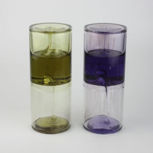 Könnte beinhalten: Zwei gläserne, sanduhrartige Behälter mit klarem und farbigem Flüssigkeitsinhalt. Der Behälter links hat einen grünen Deckel und grüne Flüssigkeit, der Behälter rechts einen violetten Deckel und violette Flüssigkeit.