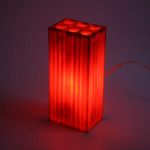 Pu&ograve; includere: Una lampada rettangolare rossa con scanalature verticali e una serie di aperture circolari sulla parte superiore. La lampada emette un bagliore rosso caldo e un cavo di alimentazione si estende dal lato. &Egrave; un apparecchio di illuminazione decorativo.