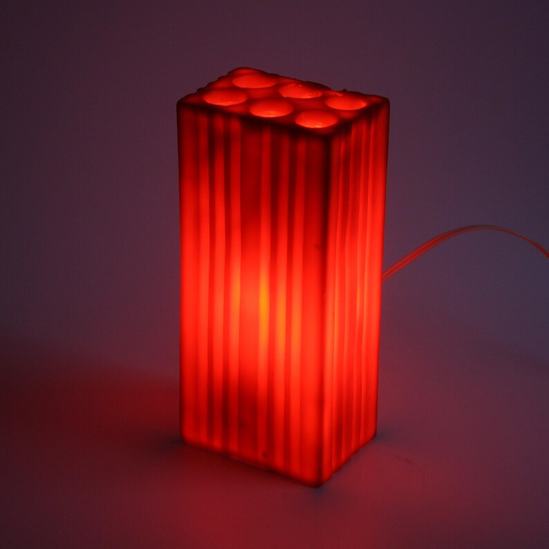 Pu&ograve; includere: Una lampada rettangolare rossa con scanalature verticali e una serie di aperture circolari sulla parte superiore. La lampada emette un bagliore rosso caldo e un cavo di alimentazione si estende dal lato. &Egrave; un apparecchio di illuminazione decorativo.