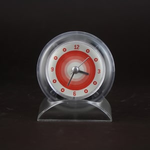 Puede incluir: Un despertador transparente de estilo retro con una esfera circular con círculos concéntricos rojos y blancos. El reloj tiene una carcasa de plástico transparente y una base curva. Las manecillas son plateadas y los números están marcados con puntos rojos.