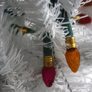 May include: Close-up of a white Christmas tree with two decorative lights. One light is a deep pink colour, and the other is orange. The lights have gold-coloured bases and green wiring.