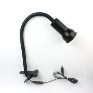 Könnte beinhalten: Eine schwarze Schreibtischlampe mit Schwanenhals und Klemmfuß. Die Lampe hat einen flexiblen, geriffelten Hals und einen konisch geformten Kopf. Das Netzkabel ist schwarz mit einem zweipoligen Stecker. Die Lampe ist zum Anklemmen an einen Schreibtisch oder eine andere Oberfläche konzipiert.