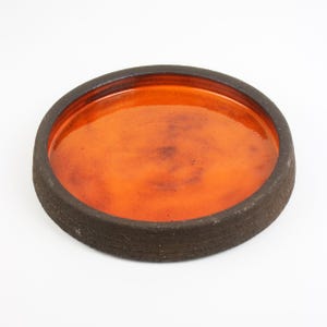 Puede incluir: Un plato redondo de cerámica con un borde marrón oscuro texturizado y un interior naranja brillante. El plato está centrado sobre un fondo blanco, mostrando su diseño simple pero llamativo. Esta pieza de cerámica hecha a mano es ideal para servir o exhibir.