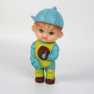May include: A vintage figurine of a child dressed as a baseball player. The figure wears a blue cap and uniform with a yellow vest. It holds a brown glove and brown shoes. The figurine has a sweet expression.