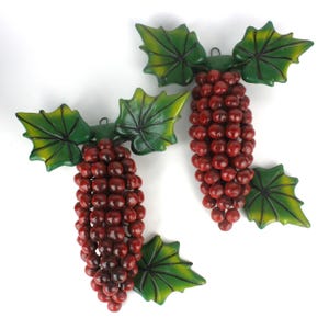 May include: Two decorative clusters of red grapes with green and yellow leaves. The grapes are round and clustered together, with a glossy finish. The leaves have a detailed veining pattern.