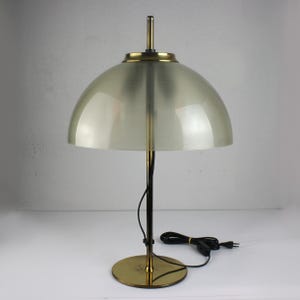 May include: A vintage table lamp with a dome-shaped shade. The lamp features a brass base and accents, a black stem, and a cord with a plug. The shade is a light, translucent color. The lamp is designed for indoor use.