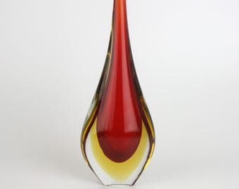 May include: A tall, teardrop-shaped glass vase with a vibrant color scheme. The vase features a striking combination of red, yellow, and clear glass, creating a layered effect. The vase has a narrow neck and a wide, rounded base.