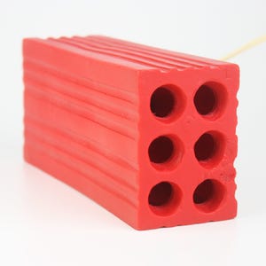 Pu&ograve; includere: Un oggetto rosso vivo a forma di mattone con sei fori circolari e scanalature orizzontali. Il mattone ha una superficie strutturata ed &egrave; realizzato in un materiale solido. Potrebbe essere un giocattolo o un oggetto decorativo.