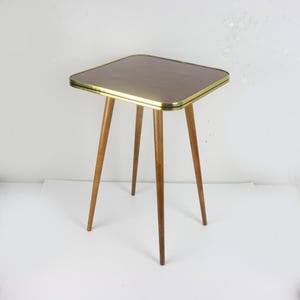 Op de afbeelding: Een vierkante bijzettafel met een donkerbruin houtnerfblad en een metalen goudkleurige rand. De tafel heeft vier slanke, taps toelopende houten poten die vanuit het midden naar buiten staan en zo een stabiele basis vormen voor het blad.