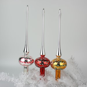 May include: Three vintage Christmas tree toppers with silver spires and round, painted glass bases. The bases are red, gold, and silver, each with a different leaf design. The toppers are displayed on a white, artificial tree branch.