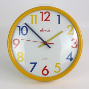 Pode incluir: Um relógio de parede redondo com moldura amarela e mostrador branco. O relógio apresenta números e ponteiros multicoloridos, com o texto "eli-vas" e "QUARTZ" impresso no mostrador. Os ponteiros indicam as horas.