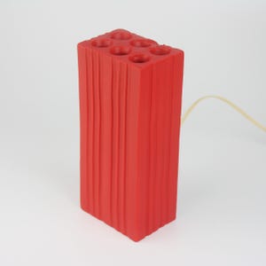 Pu&ograve; includere: Una lampada rettangolare rosso vivo con un design a forma di mattone. La parte superiore della lampada presenta sei aperture circolari. I lati della lampada presentano scanalature verticali. Un cavo giallo si estende dalla parte posteriore della lampada.