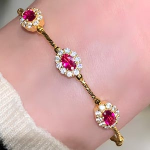 May include: A gold bracelet with three red, oval gemstones encircled by small, clear stones. The bracelet's gold chain has a textured pattern. The text "Jewelrafine USA" is visible.