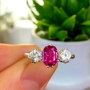 May include: A silver ring with a central oval-cut ruby and two round diamonds. The ruby is a vivid pink, and the diamonds are clear. The ring is displayed against a backdrop of green plants, with the focus on the gemstones.
