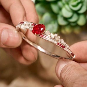 Puede incluir: Una pulsera de plata con un gran rubí ovalado y piedras rojas y claras más pequeñas. La pulsera se sostiene entre dos manos, con una planta verde borrosa en el fondo. Es una joya.