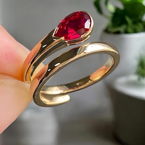 Op de afbeelding: Een gouden ring met een peervormige, gefacetteerde rode edelsteen. De band van de ring buigt om de vinger, met de edelsteen aan de bovenkant. De ring wordt tegen een vage achtergrond met een plant gehouden.
