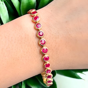 Puede incluir: Un brazalete dorado con una serie de gemas rojas vibrantes. Cada gema redonda está engastada en un bisel dorado. El brazalete se muestra sobre un fondo de follaje verde.