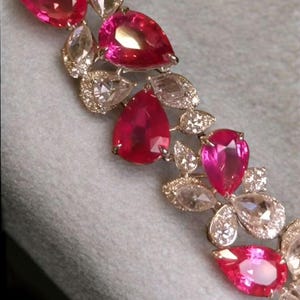 Könnte beinhalten: Nahaufnahme eines funkelnden Armbands mit tropfenförmigen, leuchtend rosa Edelsteinen und klaren, rund geschliffenen Diamanten. Die Edelsteine sind in einem silbernen oder weißgoldenen Metall gefasst und schaffen ein luxuriöses und elegantes Design.