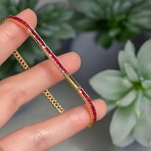 Pode incluir: Uma pulseira dourada com uma linha de pedras preciosas vermelhas de corte quadrado. A pulseira é segurada entre os dedos, mostrando o design delicado e a cor vibrante das rubis. O fecho também é visível.