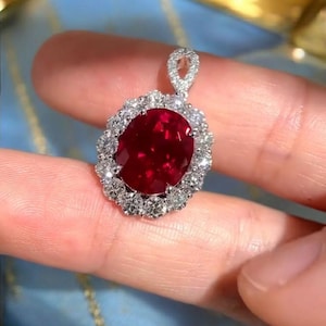 May include: A pendant featuring a large, oval-cut ruby surrounded by a halo of small, sparkling diamonds. The ruby is a deep red color, and the diamonds are clear and bright. The pendant is set in a silver or white gold setting.