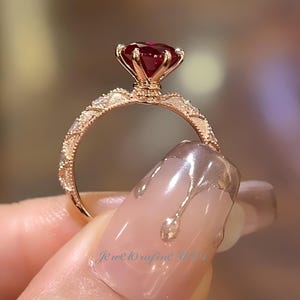 May include: A rose gold ring featuring a large, faceted, deep red gemstone. The band is intricately detailed with small embellishments. The ring is held up, showcasing the design and the gemstone's brilliance. The text "Jewelrafine World" is visible.