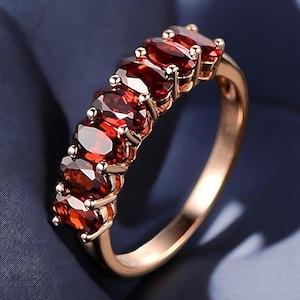 Può includere: Un anello in oro rosa con una serie di pietre preziose rosso scuro a taglio ovale. Le pietre sono incastonate vicine, creando una linea continua sulla parte superiore dell'anello. L'anello è mostrato su uno sfondo blu scuro.