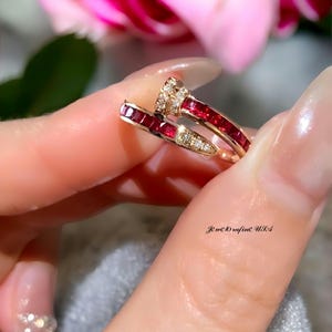 Puede incluir: Dos anillos de oro con piedras preciosas rojas y transparentes. Un anillo es una banda de piedras preciosas rojas, y el otro tiene una banda de piedras preciosas rojas y un grupo de piedras preciosas transparentes. Los anillos son sostenidos por una persona.