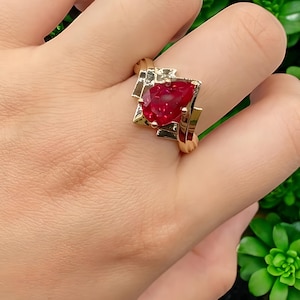 Peut inclure: Une bague dorée ornée d'une grosse pierre précieuse rouge foncé en forme de poire. La pierre est sertie dans un cadre géométrique en métal doré et argenté. La bague est portée sur un doigt, sur fond vert.