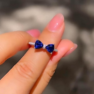 May include: A silver ring with a bow design, set with two deep blue, triangular gemstones. The bow's centre has a small, clear stone. The ring band is encrusted with small, clear stones.