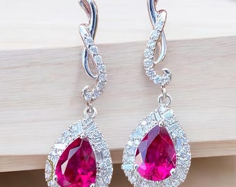May include: A pair of drop earrings with a silver-coloured design. Each earring has a large, pear-shaped, red gemstone, surrounded by smaller, clear gemstones. The earrings have a swirling design at the top.