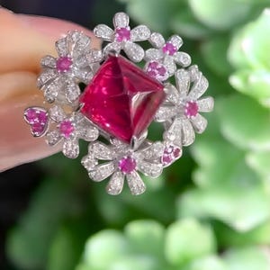 May include: A silver ring with a large, square-cut ruby gemstone at its centre. The ruby is surrounded by a floral design of small, white, diamond-set flowers with pink ruby centres. The ring is set against a blurred green background.